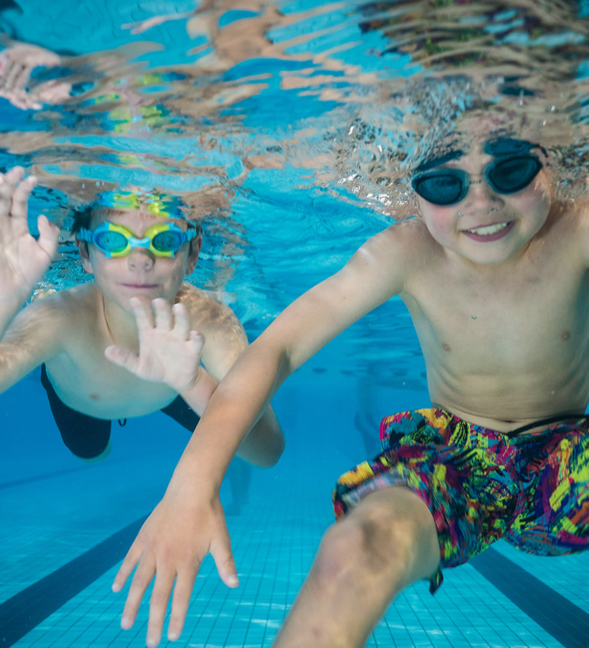 Thumbnail image. Two young children wearing swimming goggles, smile at the camera underwater.