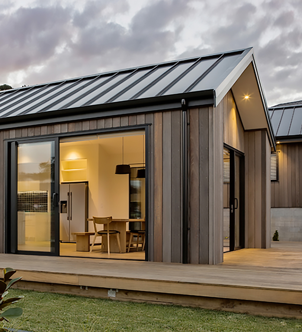 Thumbnail image. Close up shot of the outside of a David Reid Home. Parts of the interior of the house are visible through multiple glass sliding doors and windows.