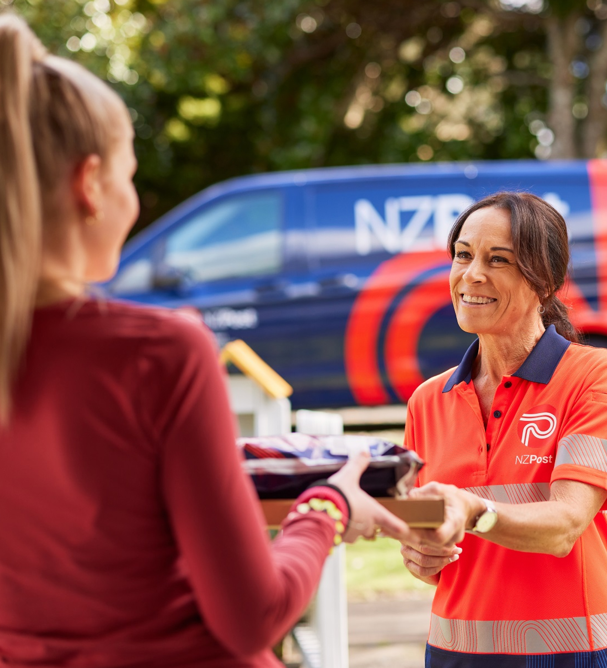 Thumbnail image. A smiling NZ Post delivery woman handing a package to the recipient outside of their house. An NZ Post van can be seen in the background.