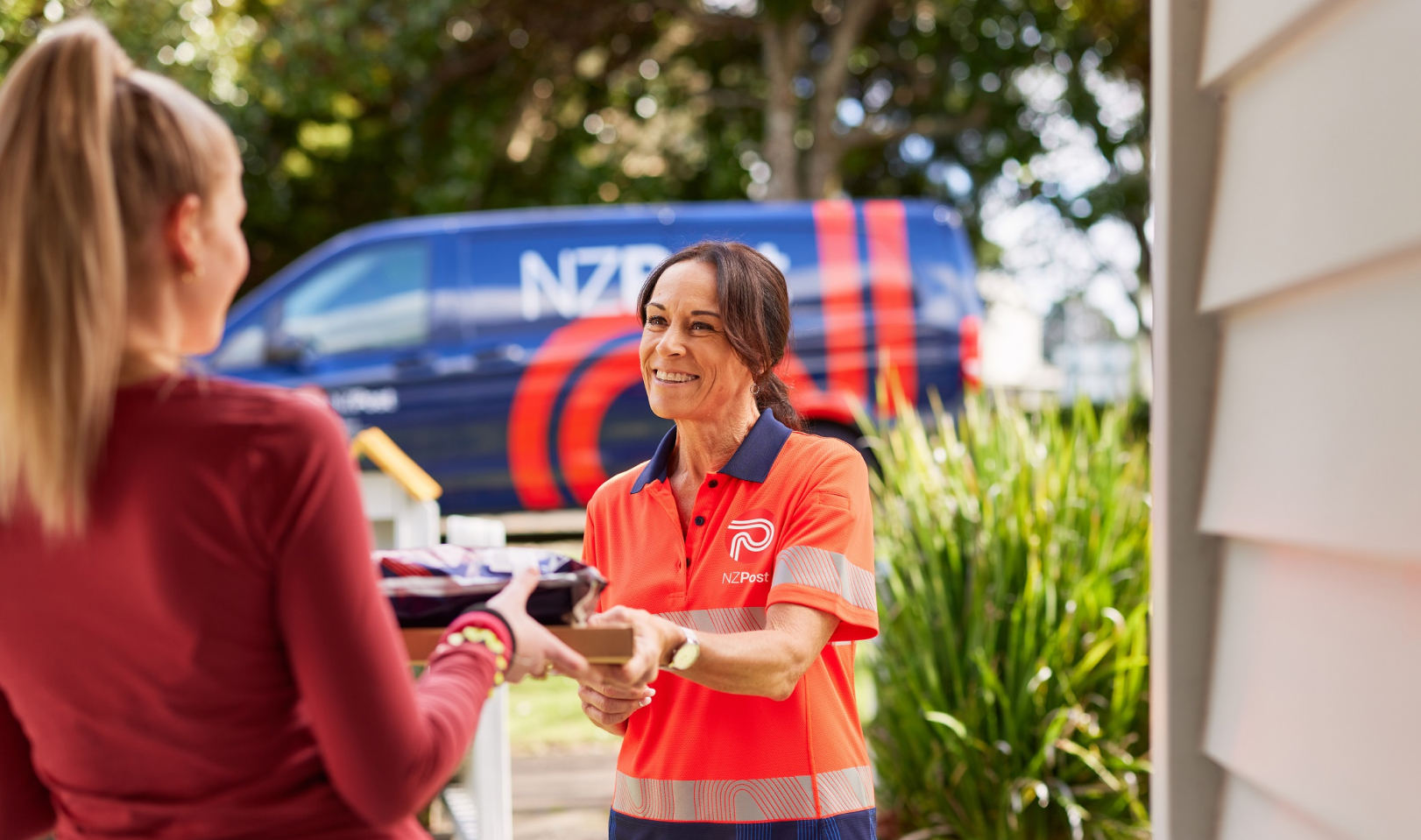 Hero image. A smiling NZ Post delivery woman handing a package to the recipient outside of their house. An NZ Post van can be seen in the background.