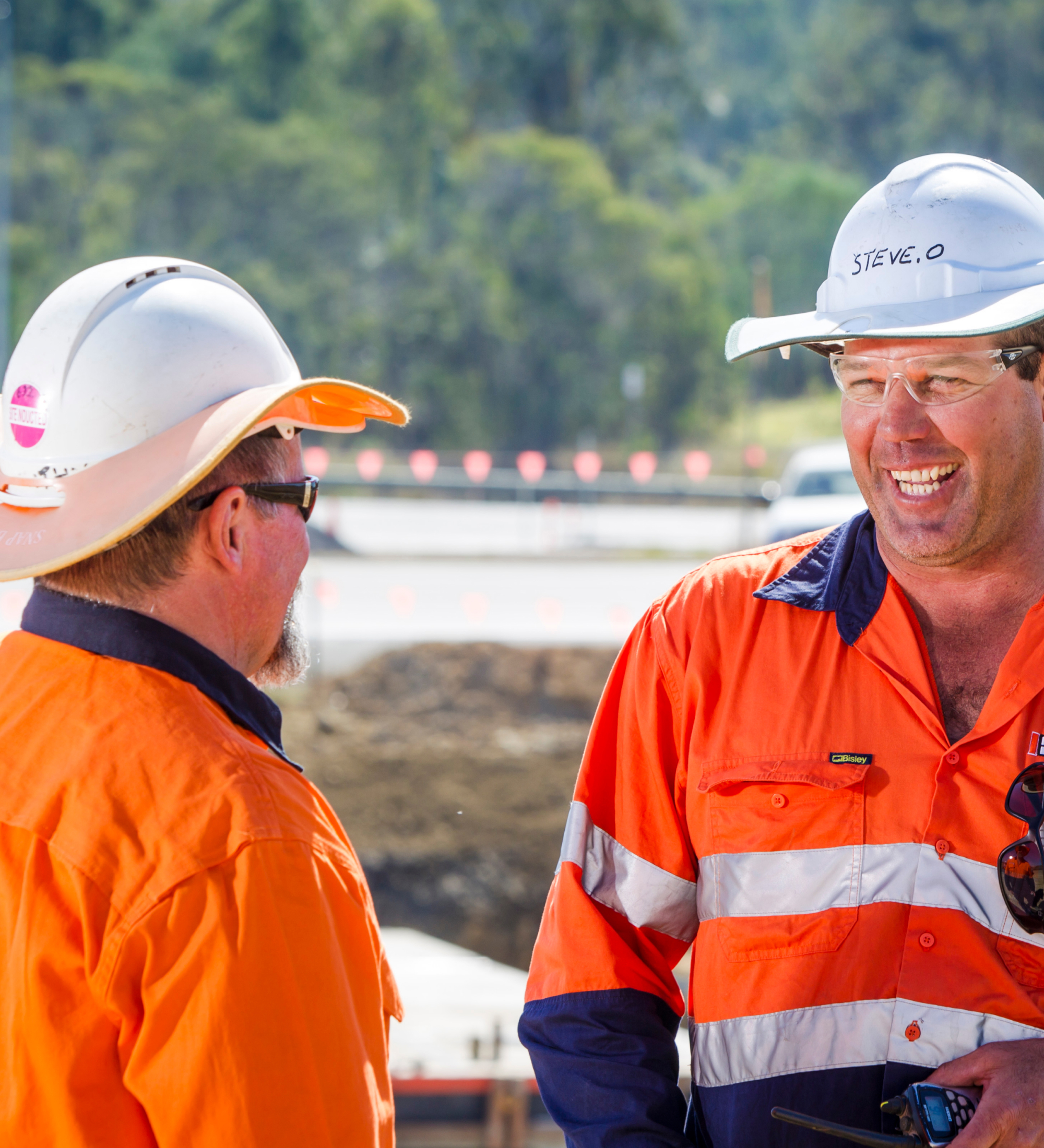 Thumbnail image. Two trades-people in orange high-visibility attire smiling and talking to one another on a building site.